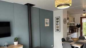 Un salon moderne doté d’un téléviseur à écran plat, d’un poêle à bois, d’un coin repas avec une table et des chaises, et d’une cuisine visible à travers une porte ouverte.