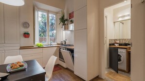 Una cocina compacta con armarios blancos, una mesa de comedor con sillas, un reloj y una ventana con plantas.