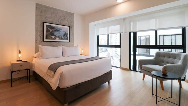 A modern bedroom with a large bed, a nightstand, a chair, and a large window with a city view. - w* | Executive 1BR Duplex in Miraflores (Miraflores)