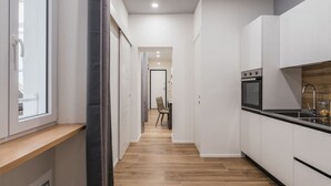 Un couloir étroit doté d’un plancher en bois, de murs blancs et d’un coin cuisine avec des armoires blanches et un comptoir noir.