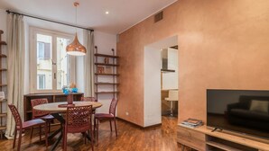 Een moderne woonkamer met een ronde houten eettafel, rode stoelen, een televisie op een houten dressoir en een boekenkast.