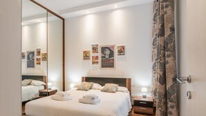 Een moderne slaapkamer met een groot bed, een houten hoofdbord en een spiegel. Er hangen kunstwerken aan de muur en een gordijn met een bladmotief.