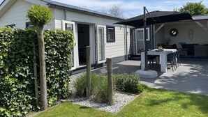 Ein modernes Haus mit einer Terrasse, einem überdachten Sitzbereich im Freien und einem Garten mit Hecke und Baum.