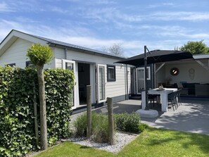 Uma casa moderna com um pátio, um espaço coberto para sentar e um jardim com um viveiro e uma árvore.
