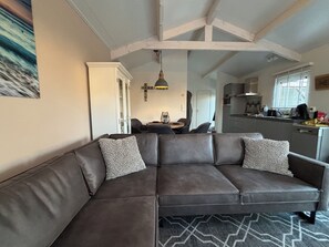Uma sala de estar moderna com um sofá seccional marrom escuro, uma área de jantar com mesa redonda e uma cozinha ao fundo.