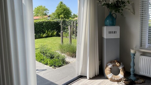 Ein Zimmer mit einer Schiebetür zum Garten, einer blauen Vase mit Blumen, einem weißen Schrank und einem Korbwägelchen.