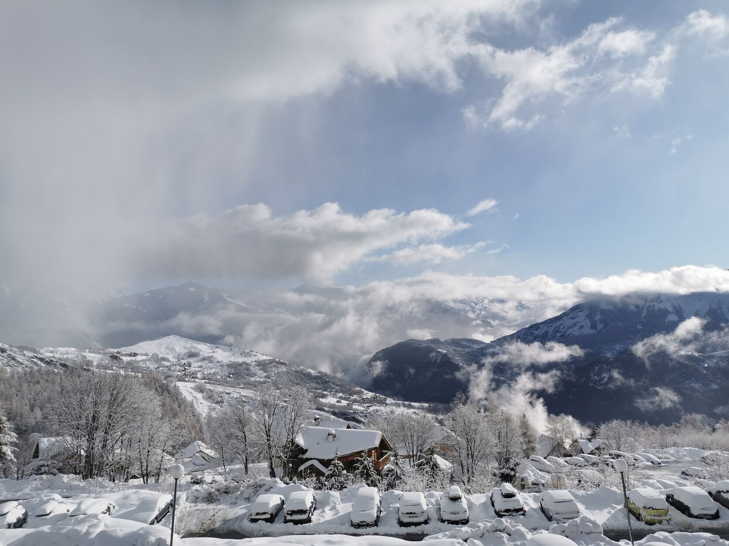 A snow-covered mountain landscape with a chalet and parked cars in the foreground.