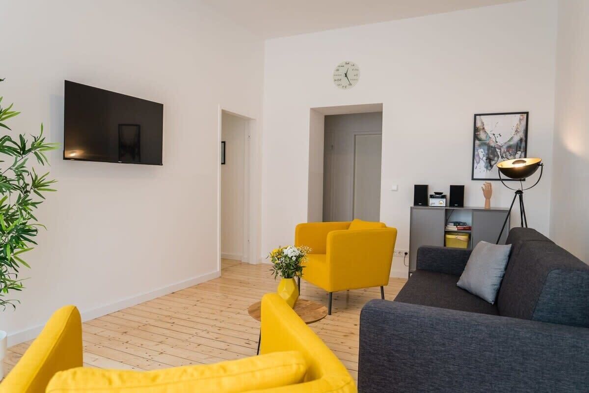 A modern living room with a flat-screen TV mounted on the wall, a sofa, a yellow armchair, and a small side table with a vase of flowers.