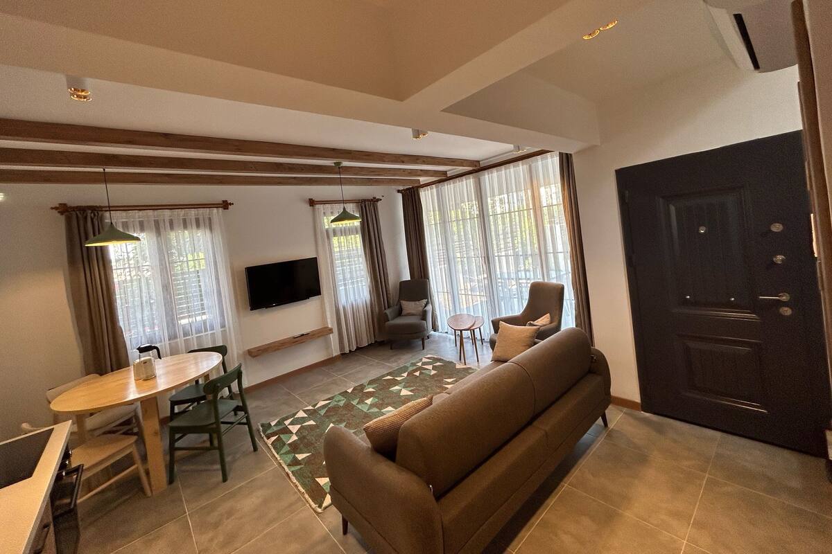 Ein modernes Wohnzimmer mit einer Couch, einem Esstisch und Stühlen. Ein Fernseher ist an der Wand befestigt und das Zimmer verfügt über große Fenster mit Vorhängen.