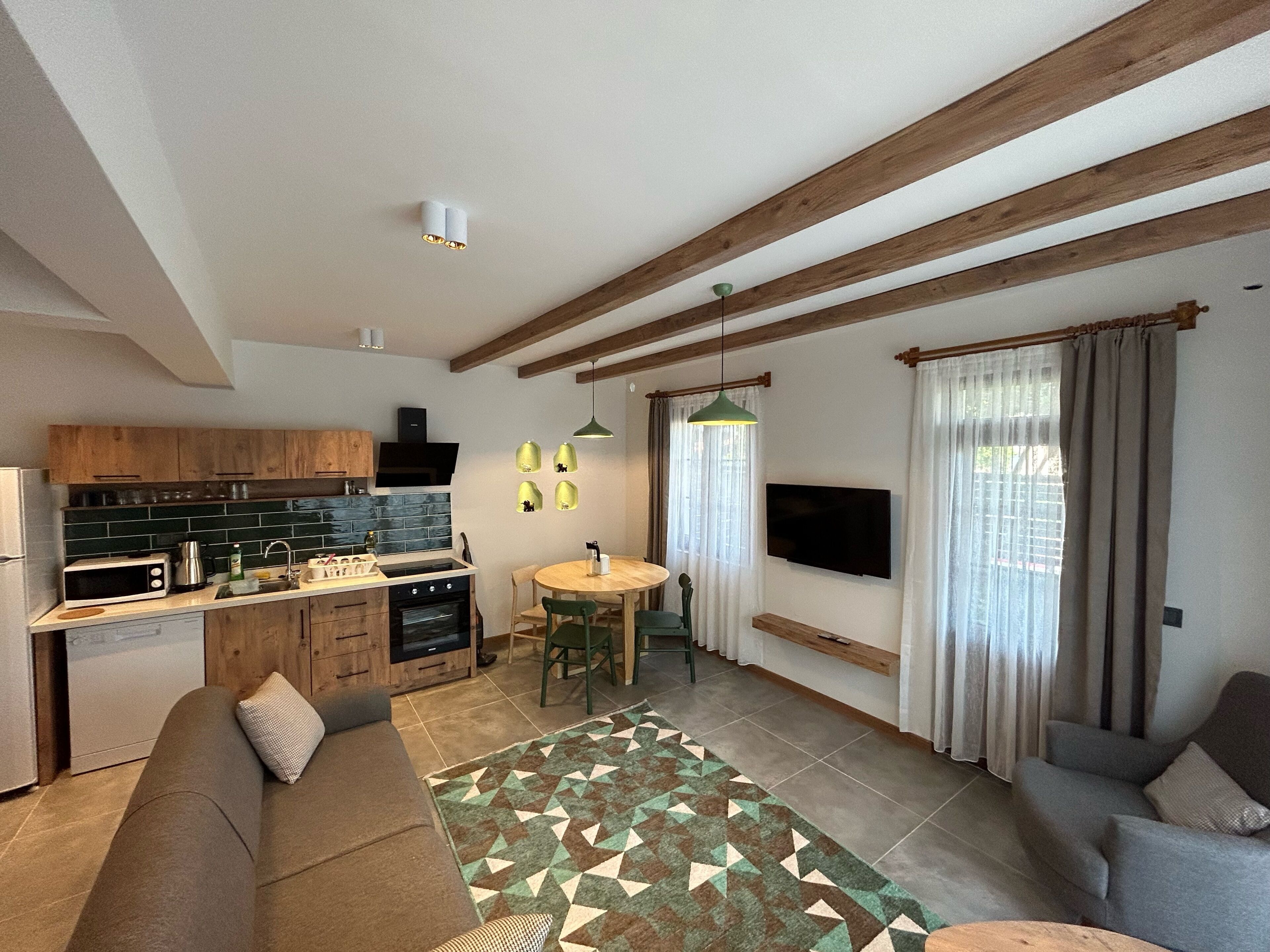 A modern kitchen with wooden cabinets, a built-in oven, and a microwave. There is a dining area with a table and chairs, and a living room with a sofa and a TV.