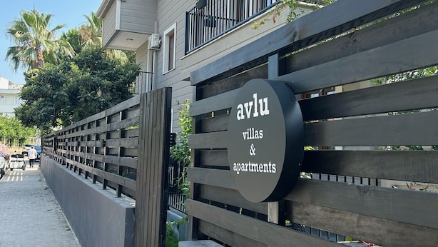 Señal de 'avlu villas & apartments' con un diseño moderno, entrada con portón y un camino despejado que lleva a la propiedad.