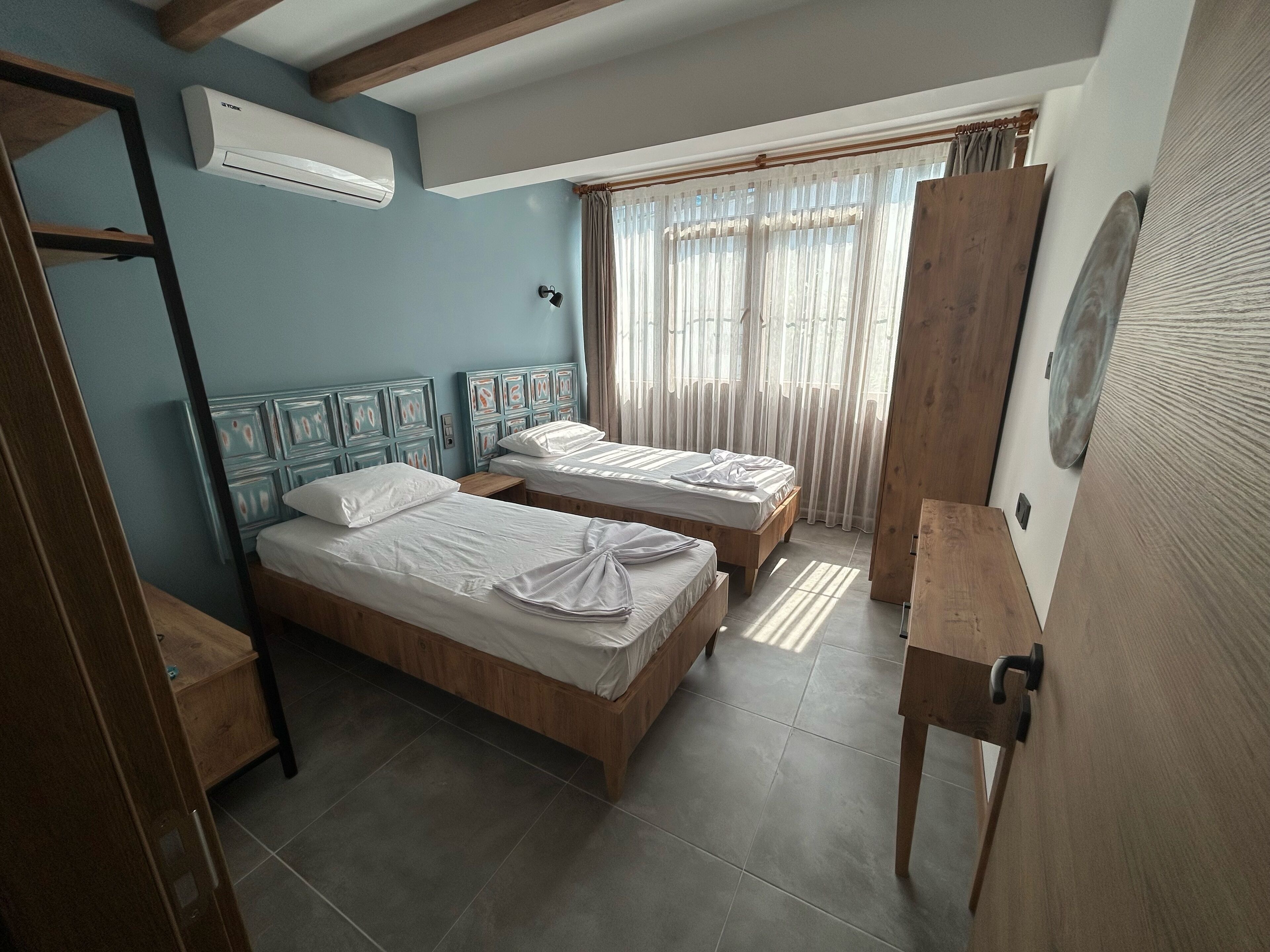 Habitación de hotel con dos camas de madera, un armario de madera, una mesita de noche pequeña, un equipo de aire acondicionado y una ventana con cortinas translúcidas.