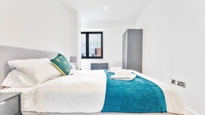 A modern hotel room with a neatly made bed, a window with bars, a grey wardrobe, and a small bedside table. - Stylish 2BR Solihull Flat, Quick Train to NEC (Birmingham)