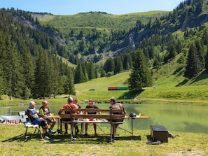 A group of people sitting at a wooden table by a lake, surrounded by a mountainous landscape.