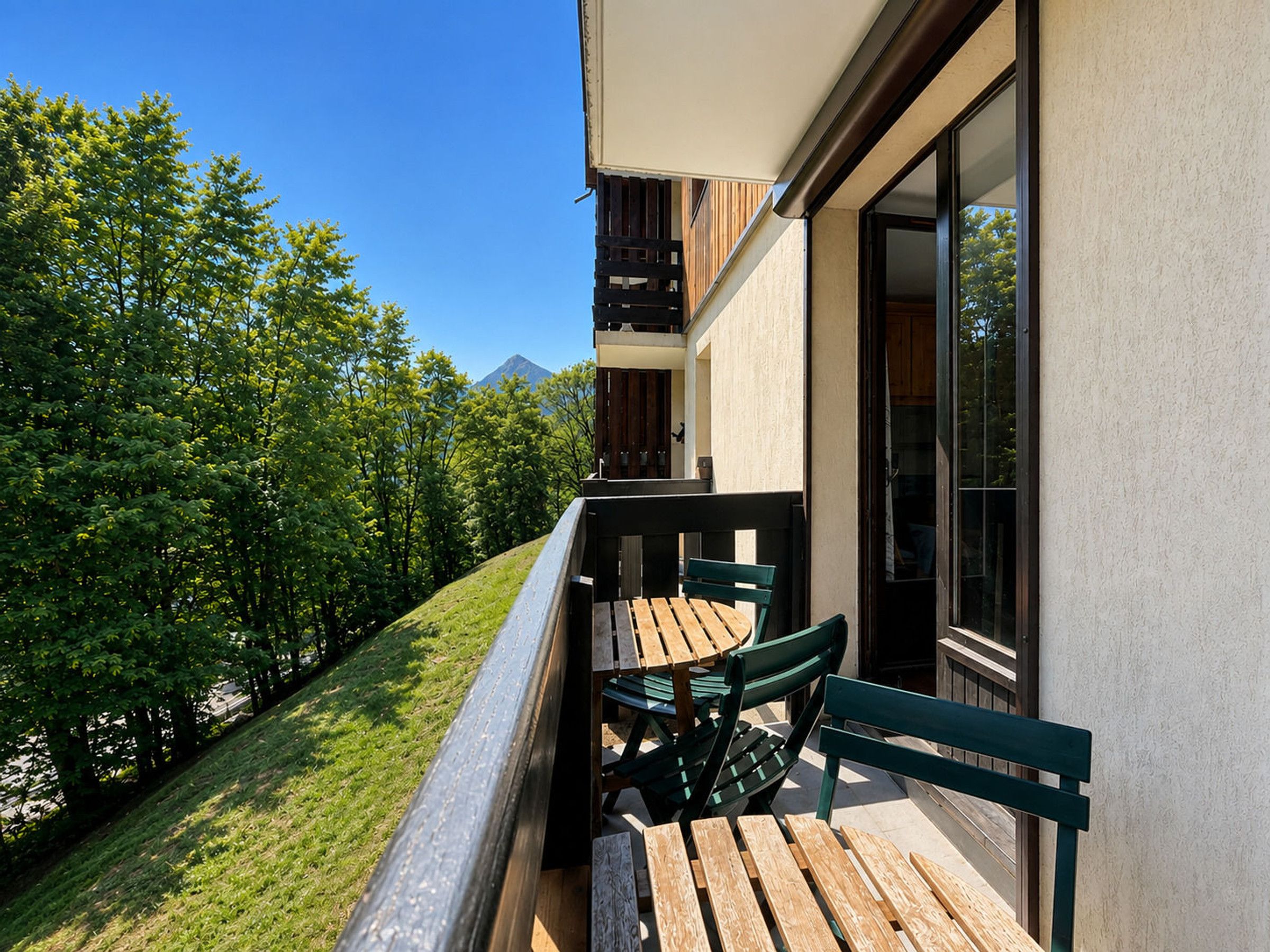 A balcony with a wooden table and chairs, overlooking a grassy area and trees.
