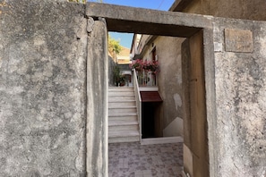 A narrow alleyway with a stone wall on one side and a tiled floor. A staircase leads to a door with a small overhang.