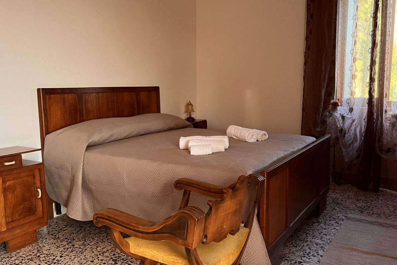 Un letto rifatto con cura, con testiera in legno, un comodino con una lampada e una sedia.