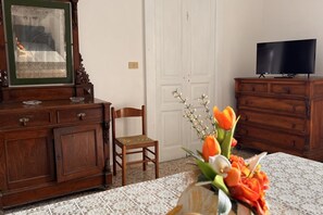 Ein Zimmer mit einem Holzkasten, einem Sessel, einem Fernseher auf einer Holzkommode und einer Tischvase mit Blumen.