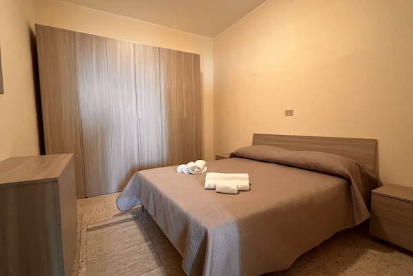 A hotel room with a large bed, a wooden wardrobe, a nightstand, and a pair of rolled towels on the bed.
