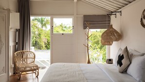 Une chambre à coucher avec un lit, une chaise en osier, un luminaire suspendu et une vue sur la verdure à l’extérieur.