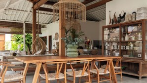 Une table à manger en bois, des chaises en osier, un lustre suspendu en rotin et une vitrine contenant des objets décoratifs.
