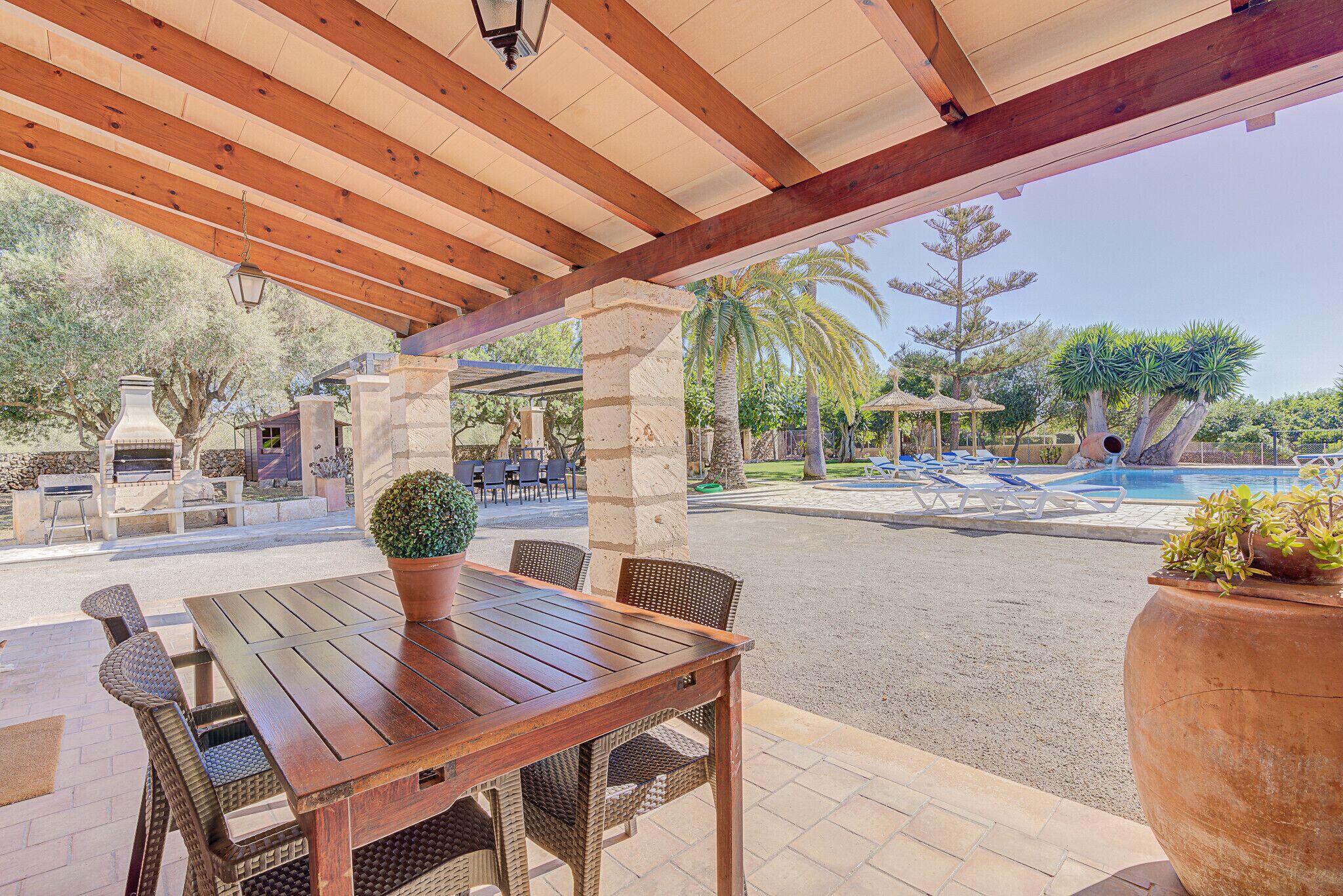 Une terrasse extérieure avec une table en bois et des chaises en osier, donnant sur un espace piscine agrémenté de chaises longues et de palmiers.