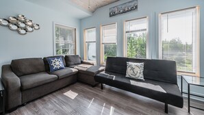 Ein modernes Wohnzimmer mit einem Ecksofa, einem Couchtisch und einem Deckenventilator. Große Fenster mit Jalousien, ein Bild an der Wand und ein dekoratives Metallwandobjekt sind zu sehen.
