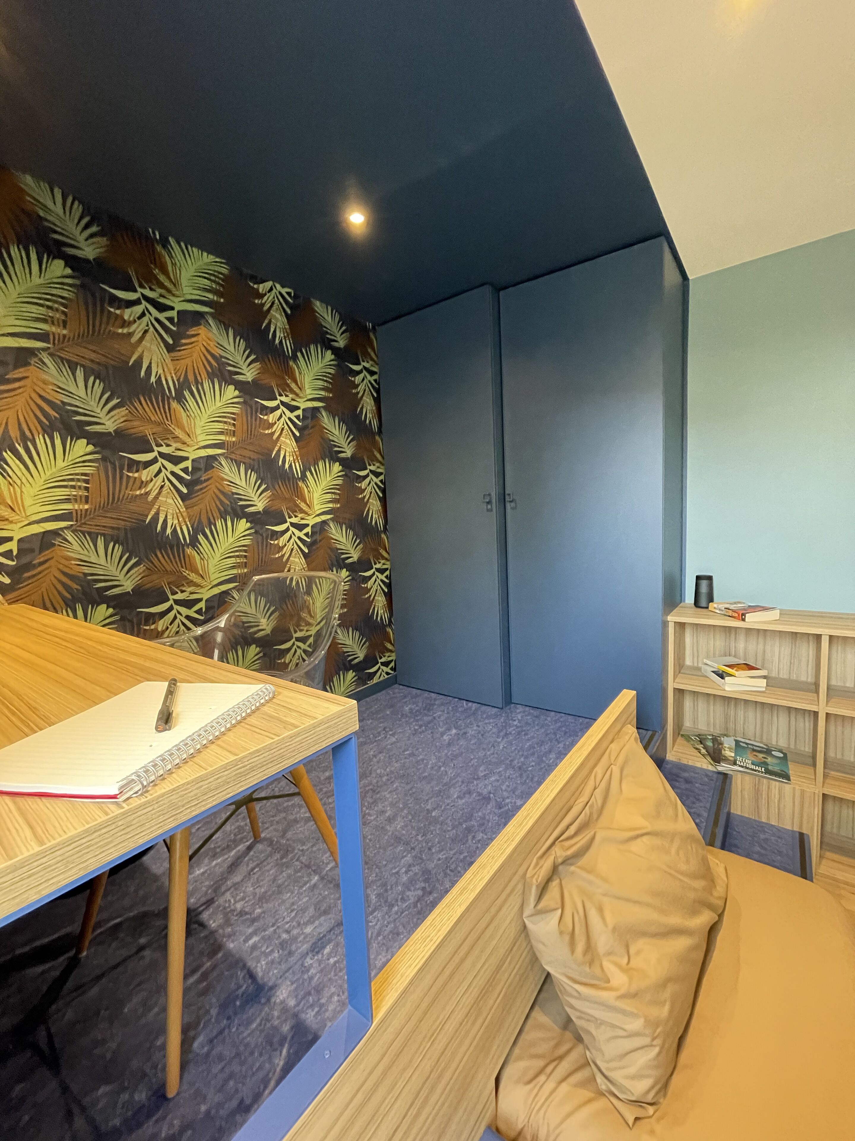 A room with a bed, a desk with a notebook, a shelf with books, and a wall with a leaf pattern.
