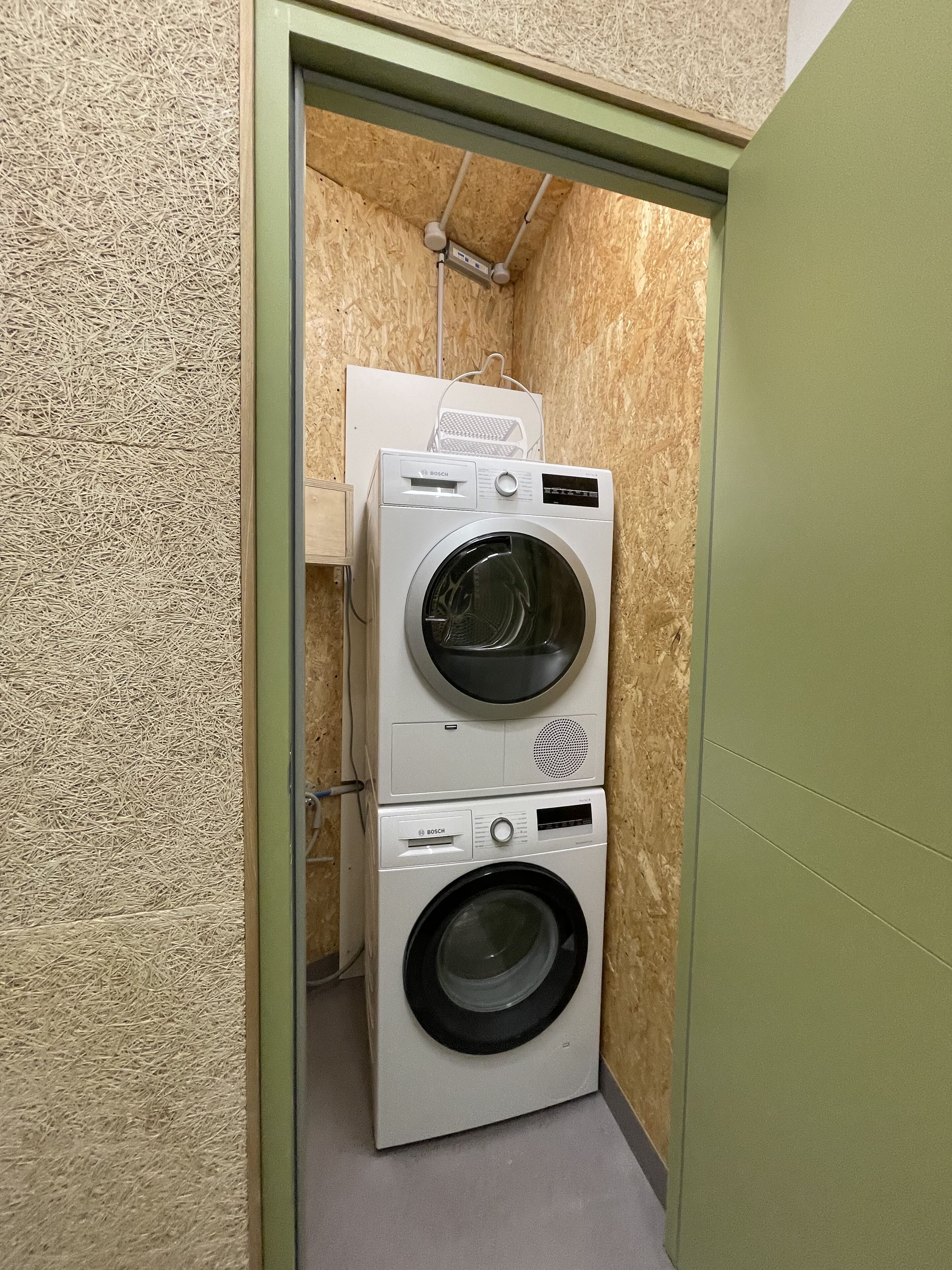 Un lavadero pequeño con dos lavadoras Bosch apiladas, un tendedero y una puerta con marco verde.