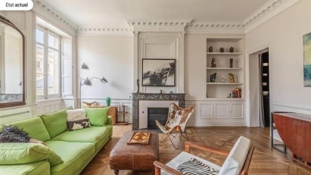Ein geräumiges Wohnzimmer mit einem grünen Sofa, einem Holztisch und einem Ledersessel. In der Mitte befindet sich ein Kamin mit Sims, und an der Wand sind Regale mit Dekorationselementen.