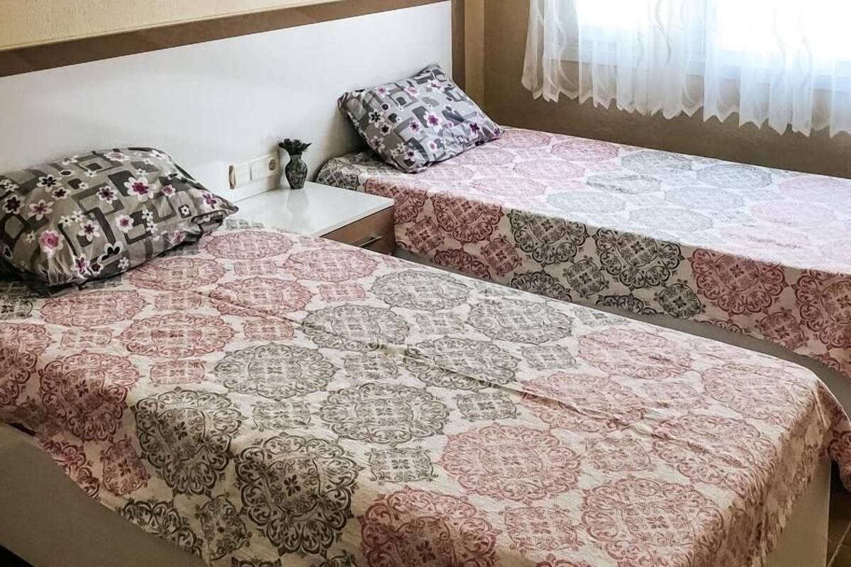 Une chambre avec deux lits, un climatiseur fixé au mur, une petite table et une fenêtre avec des rideaux légers.