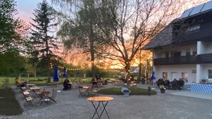 An outdoor seating area with tables and chairs, surrounded by trees and a building with a sloped roof.