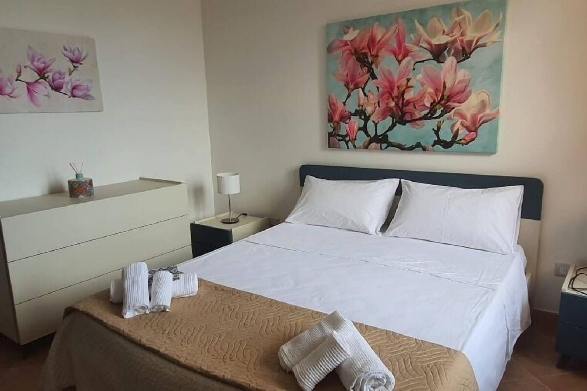 A hotel room with a bed, a nightstand, a dresser, and a painting of pink flowers on the wall.