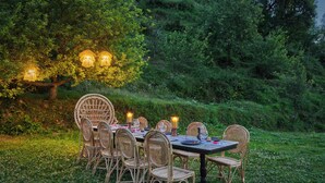 Un espacio de comedor al aire libre con sillas de mimbre y una mesa, rodeado de follaje e iluminado con luces colgantes.