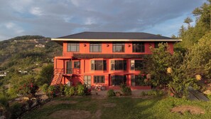 A multi-story red building with a flat roof, surrounded by greenery and hills in the background. - MINIMALIST An Eco Farmstay (Shimla)