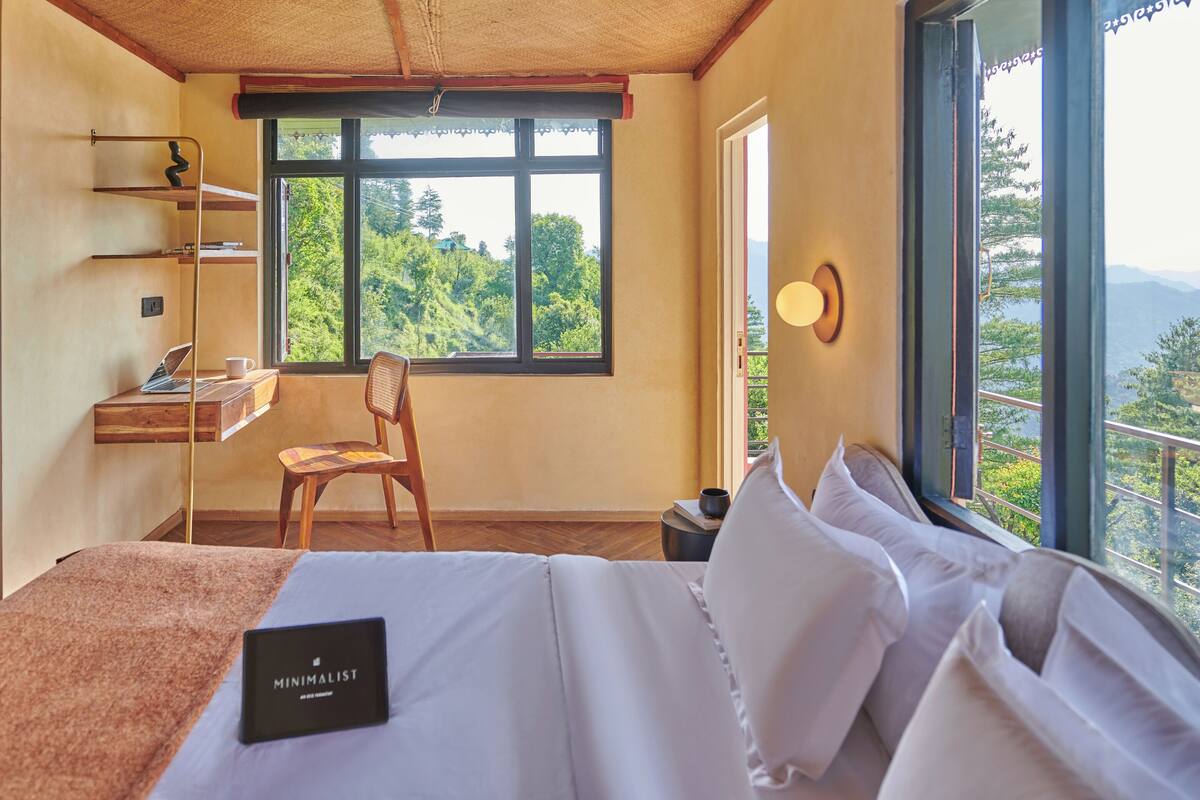 Ein Schlafzimmer mit Bett, Schreibtisch, Stuhl und Blick auf Grün durch große Fenster.