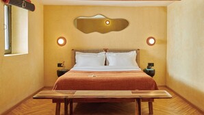 Una habitación de hotel con cama, cabecero de madera, dos mesitas de noche, una lámpara de noche, un espejo adosado a la pared y una ventana.