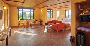 A cozy room with wooden flooring, a red sofa, a small round table, and a chair. There are two windows providing natural light and a view of greenery outside. - MINIMALIST An Eco Farmstay (Shimla)