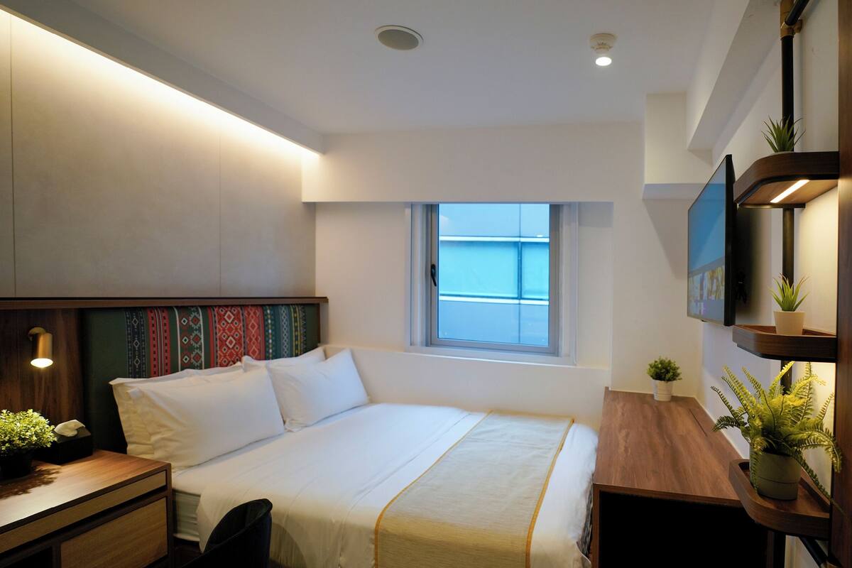 A modern hotel room with a bed, bedside table, and a small desk with a chair. There is a window providing natural light and a view of the outside.