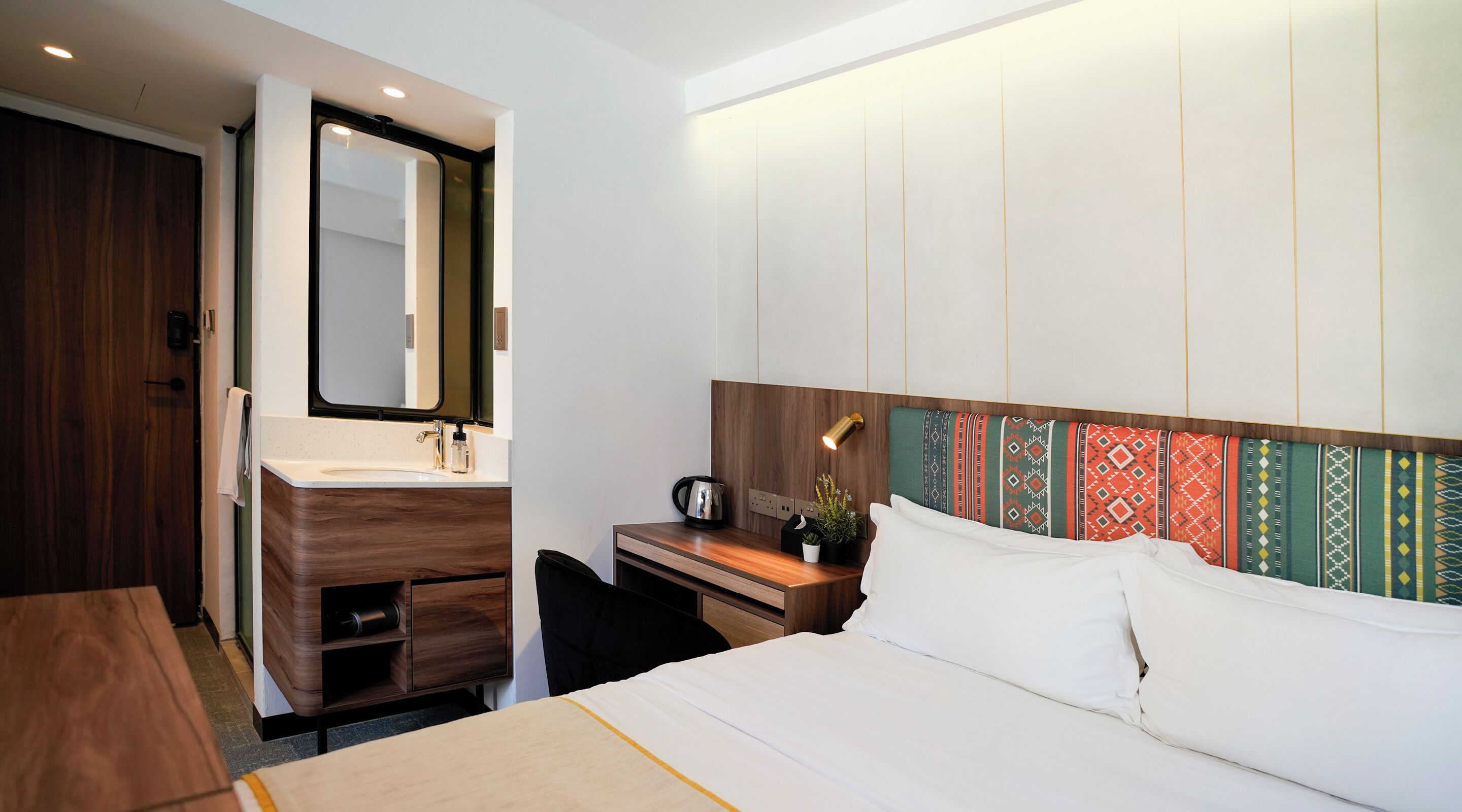 A hotel room with a wooden desk, a bed with white bedding, a large mirror, and a colorful headboard.