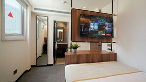 A hotel room with a bed, a desk with a plant, a TV displaying movie posters, and a mirror. - ST Signature Bugis Middle (Singapore)