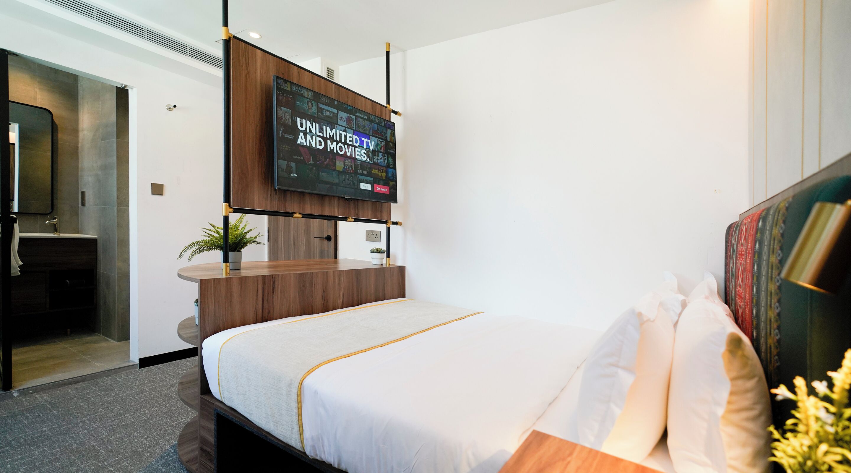 A hotel room with a bed, a TV mounted on the wall, a wooden desk, and a bathroom visible through an open door.