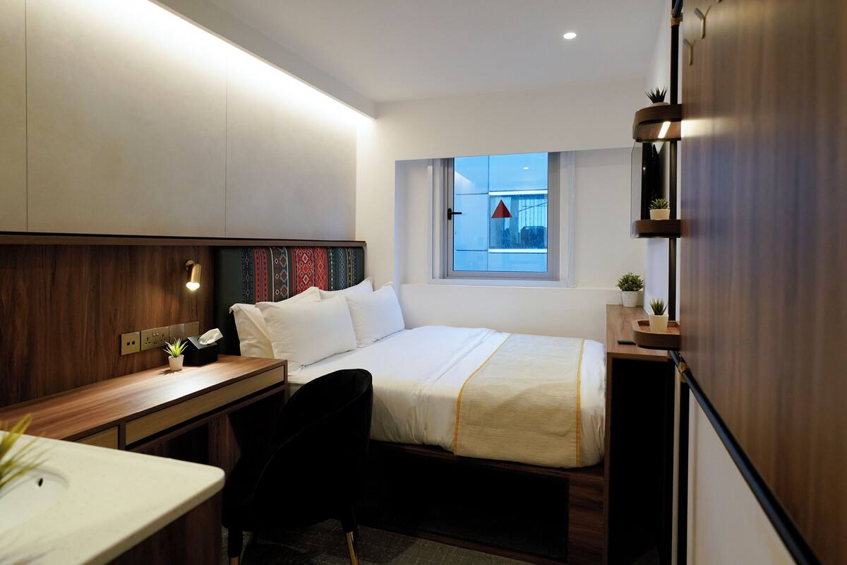 A compact hotel room with a bed, a desk, a chair, a small table, and a window with a view of the outside.