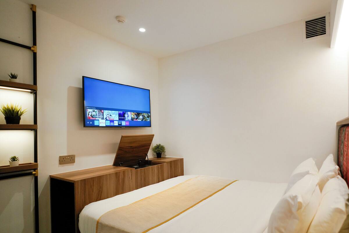 A hotel room with a bed, a TV mounted on the wall, a wooden nightstand, and a shelf with plants.