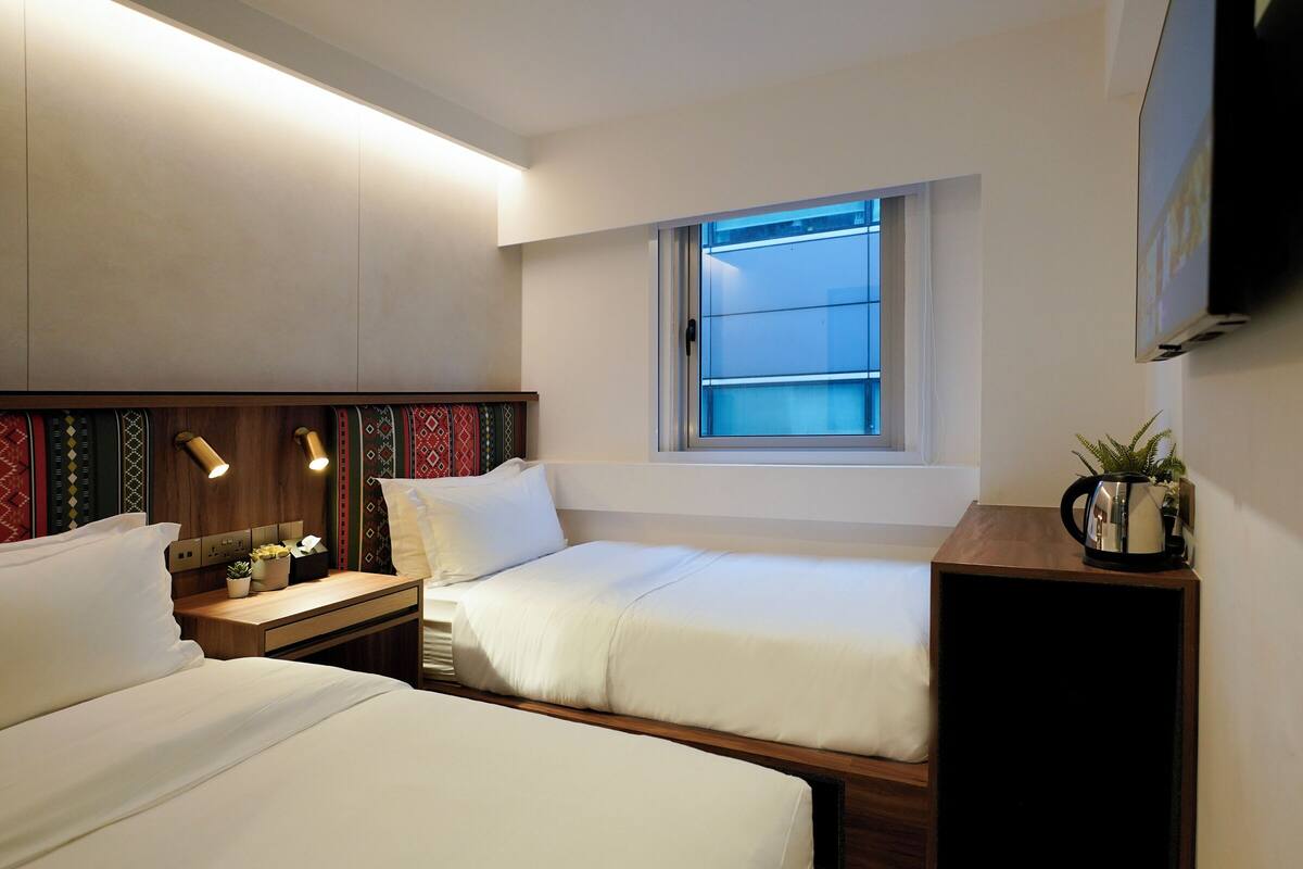 A hotel room with two beds, a window with a view, a bedside table with a lamp, and a TV mounted on the wall.
