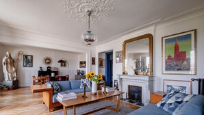 A spacious living room with a wooden floor, a sofa, a coffee table with books and a vase of sunflowers, a fireplace, and a large mirror reflecting a cityscape. - Eclectic Eminence, by Veeve (Paris)