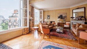 A spacious living room with a large window, wooden flooring, and a mix of seating including a sofa, chairs, and a coffee table. - Mellow Grace, by Veeve (Paris)