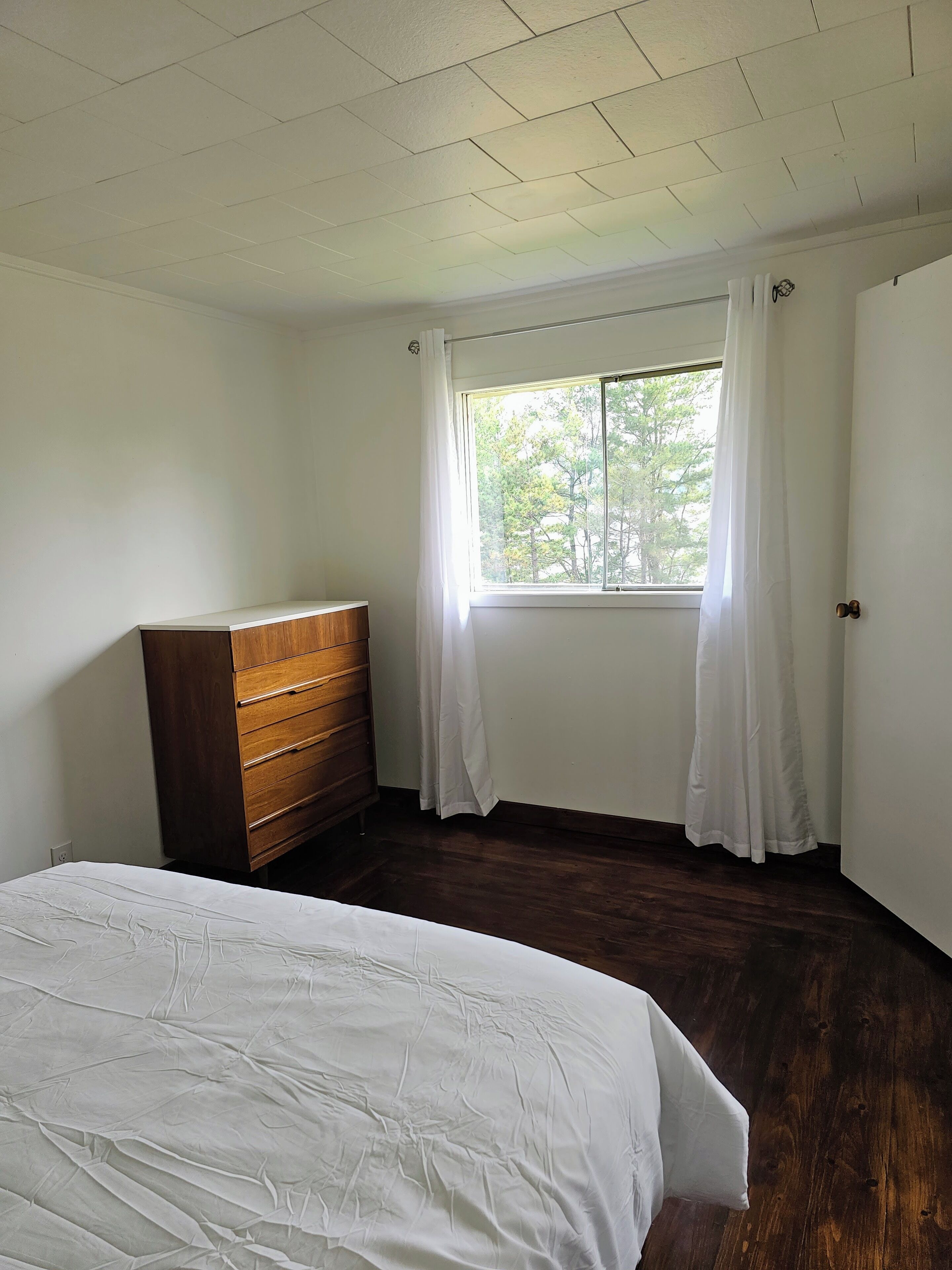 A bedroom with a wooden dresser, a bed, a window with curtains, and a door.