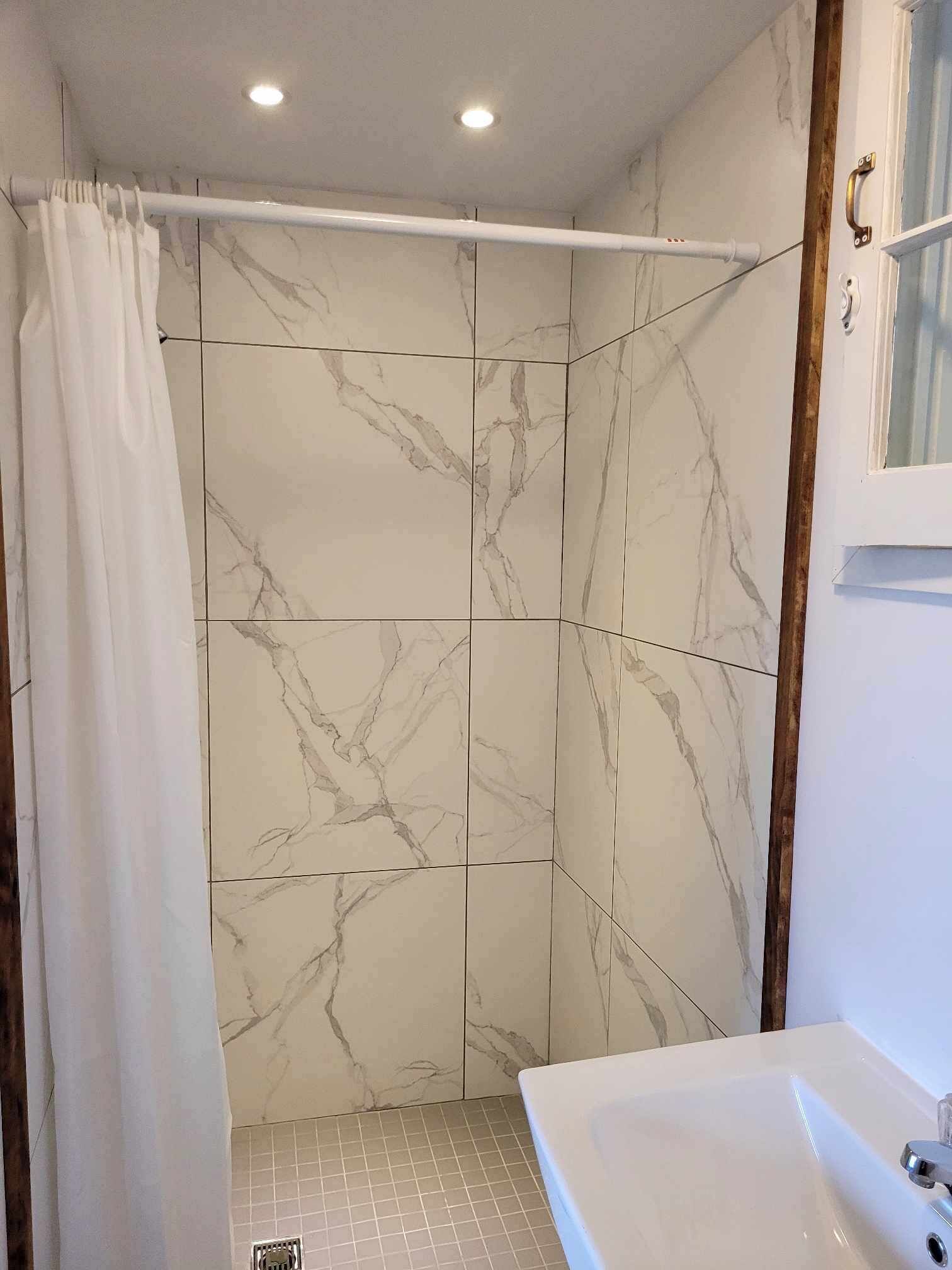 Een badkamer met marmeren tegels, een douchegordijn, een bad en een raam met houten kader.