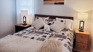 A neatly made bed with a patterned bedspread, two bedside tables with lamps, a window with a curtain, and a painting on the wall. - Mazinaw Lakeside RV Park & Campground (Cloyne)